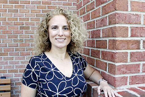 Director, Quality Systems, Christine Crone is smiling while she leans against a brick wall outside of Infinite Legacy's office.